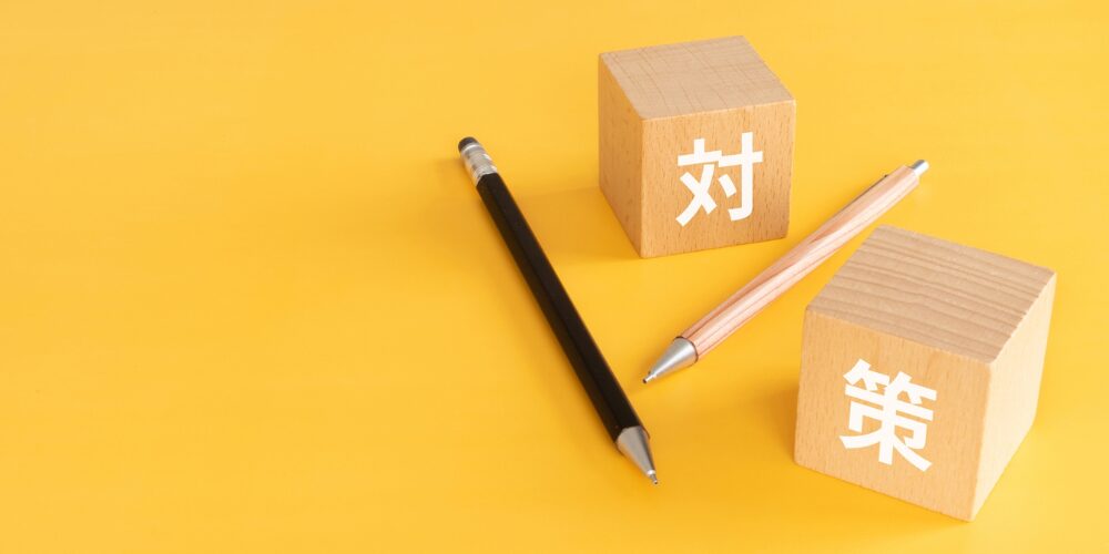 黄色い背景の上に、木製ブロックが二つ置かれ、それぞれに白文字で「対」「策」が書かれ、横に黒とナチュラルの鉛筆が斜めに配置されたシンプルなデザイン。