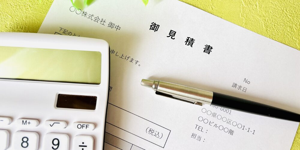 緑色の壁を背景に、白い電卓と黒いペンが乗った見積書用紙の上部を捉えた写真。用紙には『御見積書』と書かれており、ビジネスツールが並ぶ構図