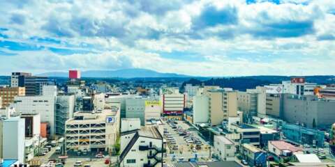 八戸市中心部を高層ビルから見下ろした街並み。広い駐車場や商業ビルが立ち並び、遠景には山々と雲多めの青空が広がる風景写真