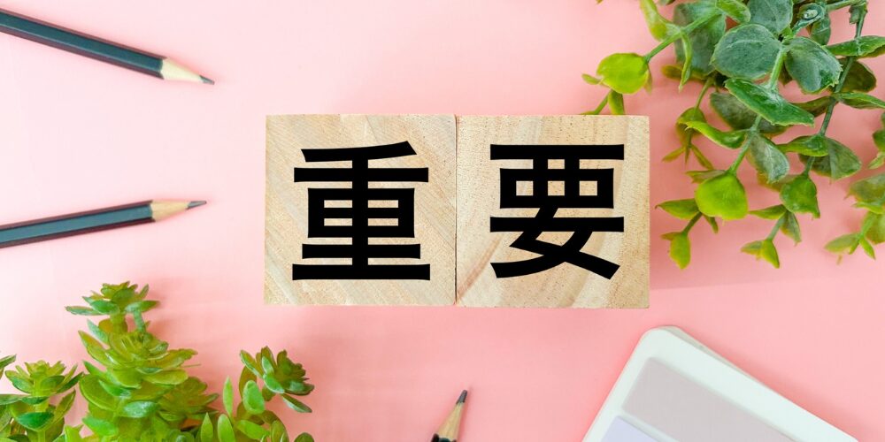 ピンクの背景に木製ブロックが並び、それぞれに黒文字で『重』『要』と書かれているクローズアップ写真。周囲にはグリーンの観葉植物と色鉛筆が配置