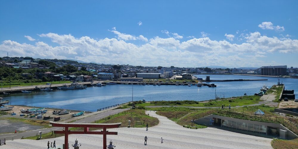 海沿いの景色。正面に赤い鳥居、その先に防波堤と漁港、市街地が広がり、青空に白い雲が浮かぶ穏やかな港の風景。
