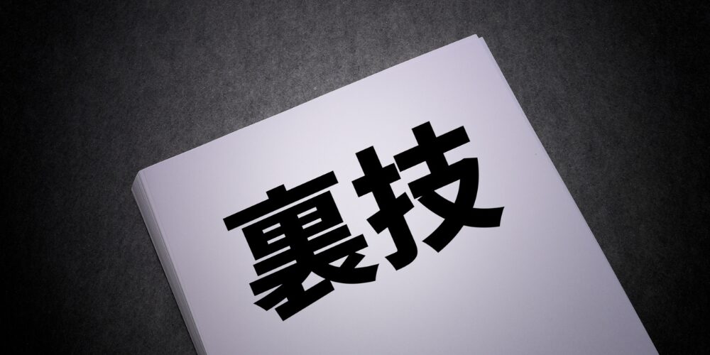 黒いテーブル上に積まれた白い紙束の上に大きな黒文字で「裏技」と印刷されたドキュメント。